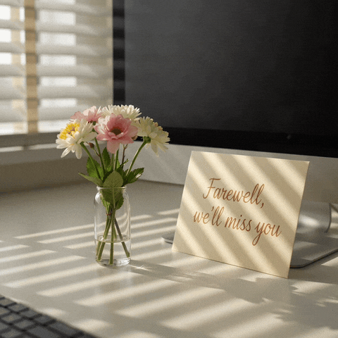 Farewell, we'll miss you handwritten card beside a vase of pink and white flowers on a sunlit office desk